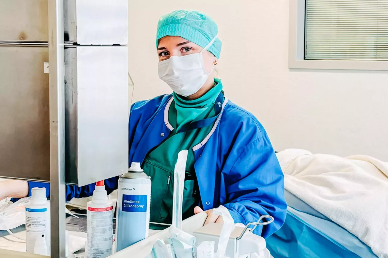 Medizinische Fachkraft in blauer Schutzkleidung und grünem Haarnetz neben einem Krankenhausbett mit blauer Bettwäsche und medizinischem Gerät.