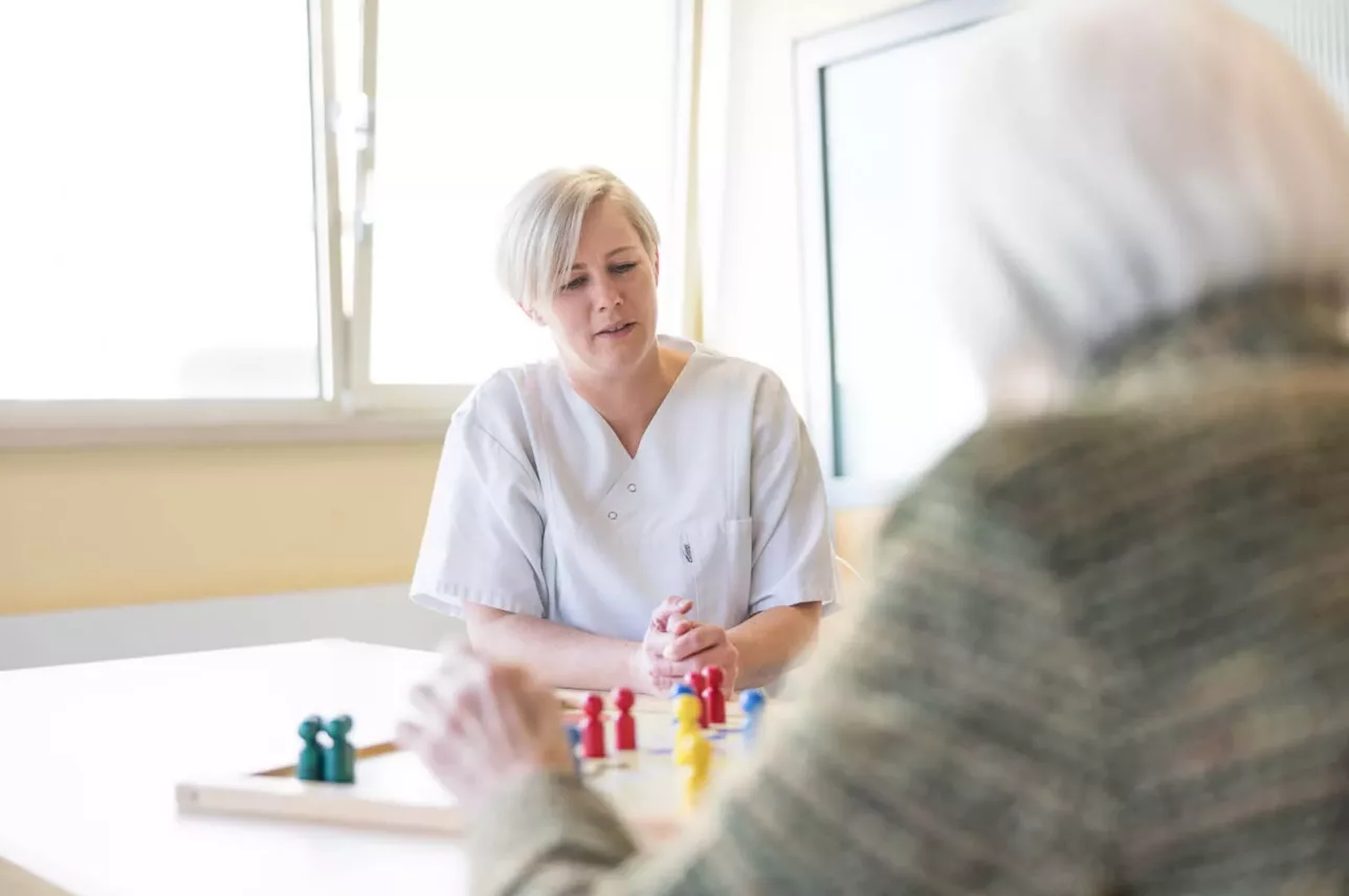 Eine Pflegekraft in weißem Kittel sitzt einer älteren Person an einem Tisch gegenüber und beobachtet sie beim Spielen eines Brettspiels mit bunten Figuren in einem hellen Raum. Die Pflegekraft sieht aufmerksam und engagiert aus.