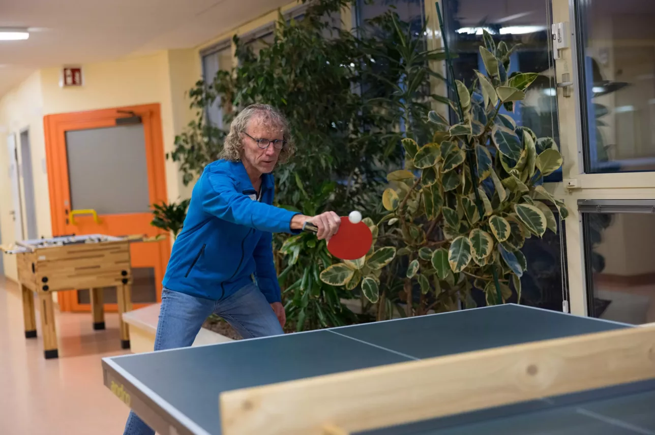 Person in blauer Jacke spielt Tischtennis, hält roten Schläger und schlägt weißen Ball über Tischtennisplatte.
