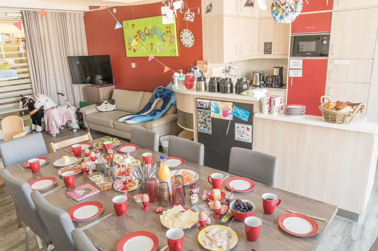 Ein hell dekorierter Raum für eine Kinderparty mit einem langen Tisch mit Snacks, Getränken und roten Tassen. Luftballons, Banner und Spielzeug sind zu sehen, mit einer Couch und einem Küchenbereich im Hintergrund.