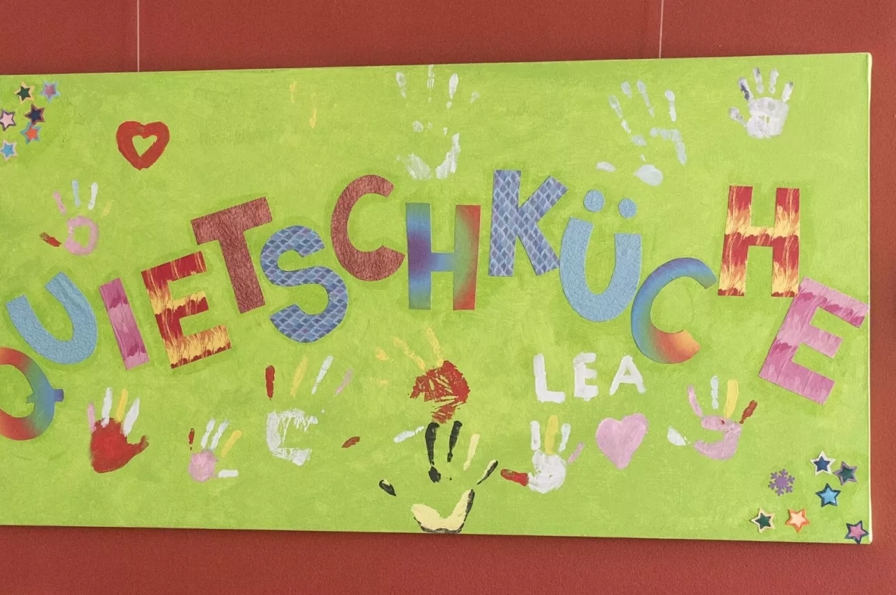 Ein buntes handgefertigtes Schild auf grünem Hintergrund mit dem Wort QUIETSCHKÜCHE in großen, bunten Buchstaben, umgeben von gemalten Handabdrücken, Herzen, Sternen und dem Namen LEA. Das Schild hängt an einer roten Wand.