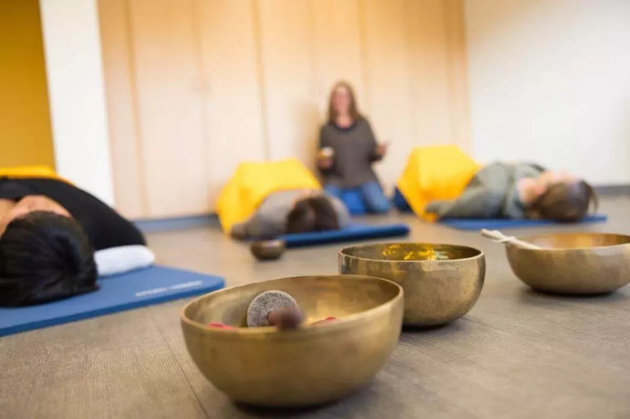 Drei Personen liegen auf Matten mit gelben Decken in einer Entspannungs- oder Meditationssitzung, während eine Person im Hintergrund sitzt. Im Vordergrund sind Klangschalen auf dem Boden aufgestellt.