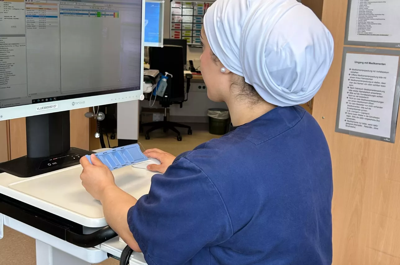 Pföegekraft steht an einem höhenverstellbaren Schreibtisch mit Computerbildschirm und hält einen Medikamentendispenser in der Hand.