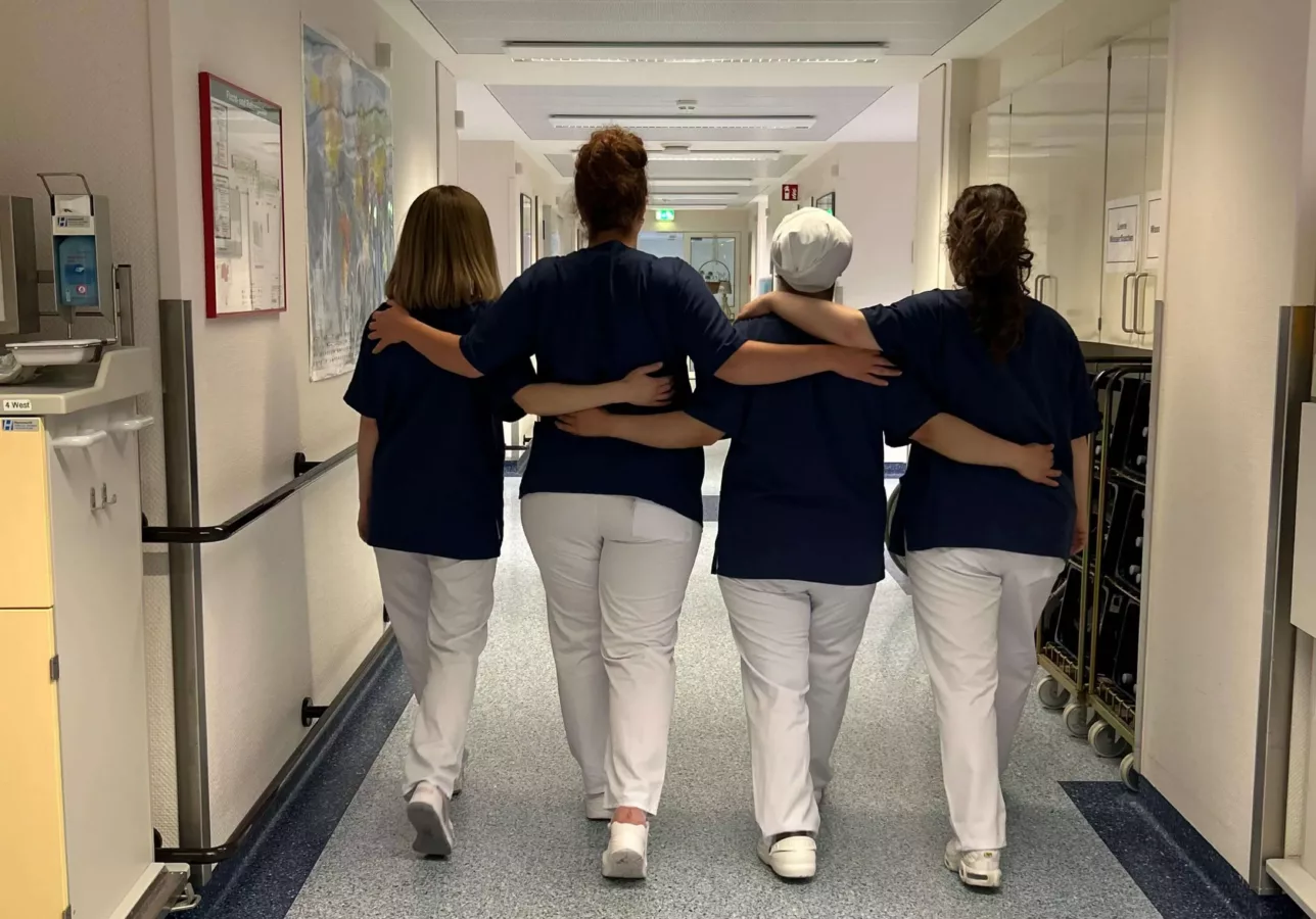 Vier Pflegekräfte gehen Arm in Arm einen Krankenhausflur entlang, Rückenansicht.