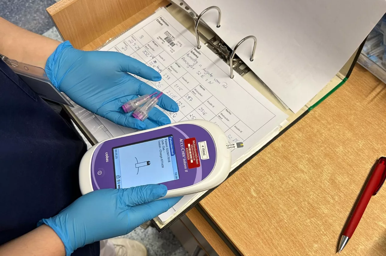 Person mit blauen Handschuhen hält medizinisches Messgerät und zwei kleine Probenbehälter über einem geöffneten Ordner mit Tabellen auf einem Schreibtisch.