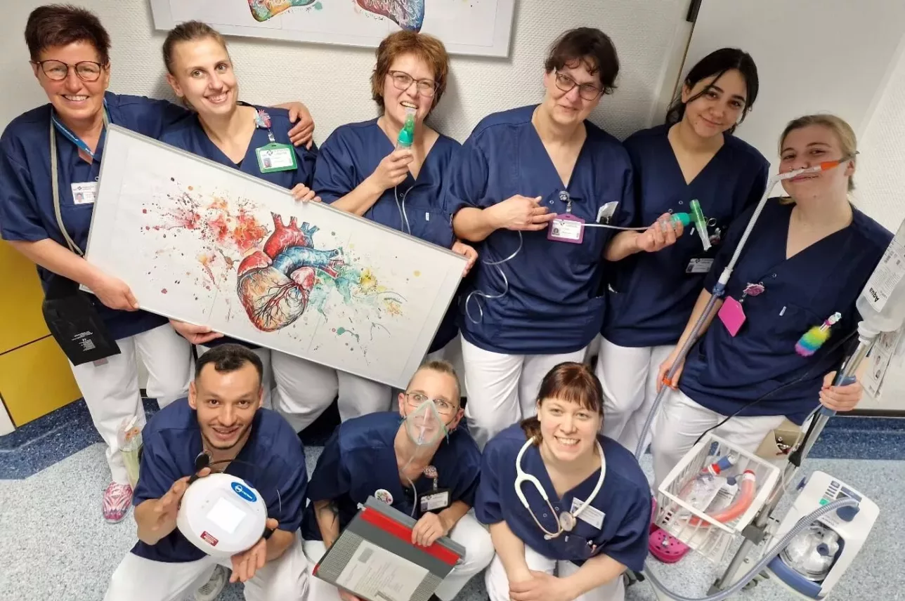 Neun Mitarbeiter des Gesundheitswesens in blauen Kitteln und weißen Hosen, die lächelnd in einer Krankenhausumgebung zusammen posieren. Einige halten medizinische Geräte und ein buntes Herz, das in einem hellen Raum steht und kniet.