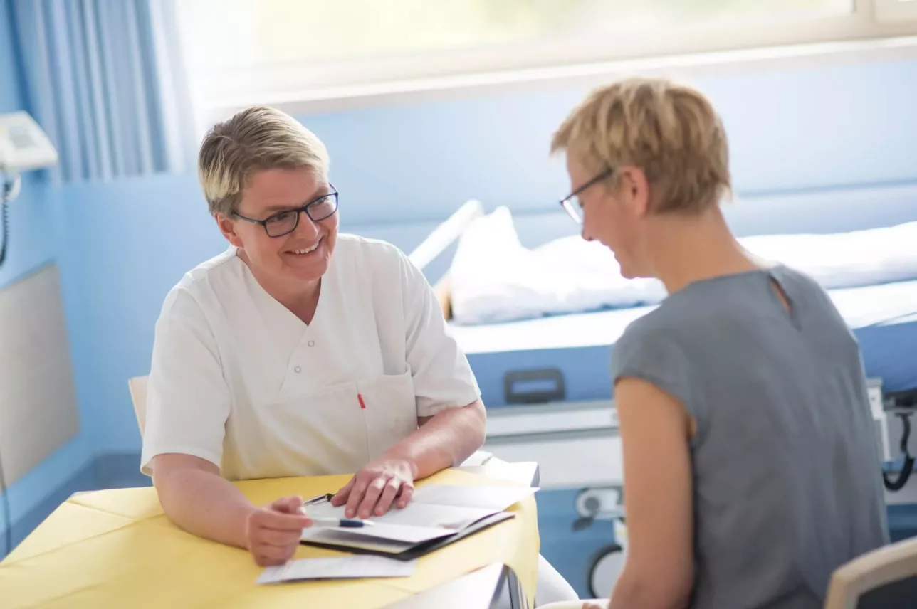 Eine medizinische Fachkraft in einem weißen Kittel lächelt, während sie mit einem Patienten in einem Krankenhauszimmer Unterlagen bespricht. Der Patient hört über einen Tisch hinweg aufmerksam zu, während im Hintergrund medizinische Geräte zu sehen sind.