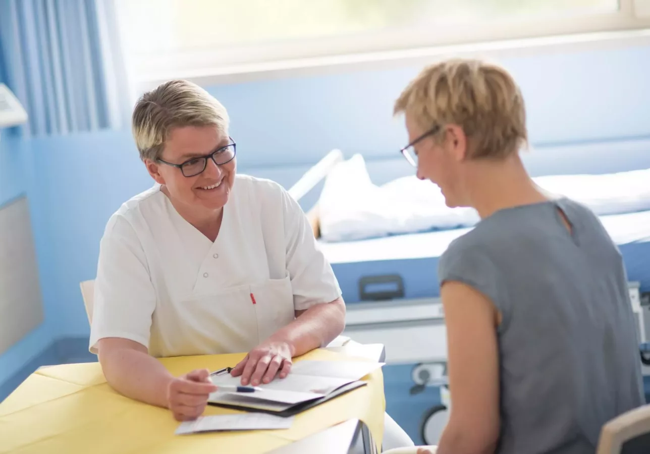 Eine medizinische Fachkraft in einem weißen Kittel lächelt, während sie mit einem Patienten in einem Krankenhauszimmer Unterlagen bespricht. Der Patient hört über einen Tisch hinweg aufmerksam zu, während im Hintergrund medizinische Geräte zu sehen sind.